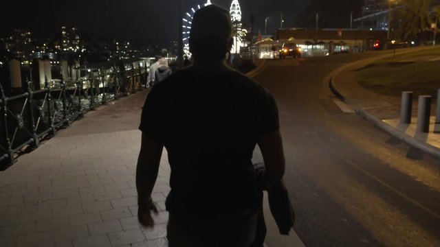 Camera Smoothly Follows Man Walking On Path Towards Theme Park at Night