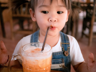 Happy little Asian girl eating thai iced tea with milk in glass at outside of the coffee cafe . Cute face.