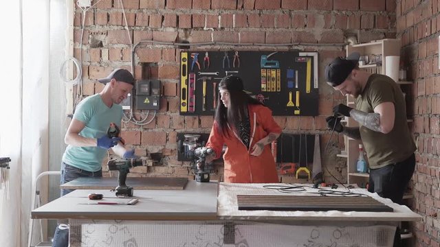 Two men and woman working in comfortable clothes and caps making furniture in the workshop. Modern people making furniture in garage. The board with the tools hanging on the wall