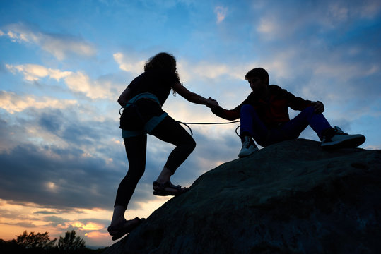 Woman is climbing on cliff. Man friendly hand helping her. Bouldering on the mountains in the evening. Silhouette of slim young woman outdoors in summer on beautiful sky background. Teamwork concept.