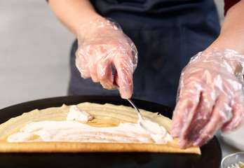 Cropped view of chef in blue apron in dish preparing process. Woman hands in one-time gloves lubricate crepe, folded up twice on black top, with creamy sauce and teaspoon. Light background. Front view