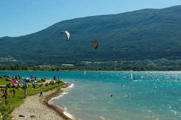 Crowded beach of the lake Le Bourget with people playing on the grass, bathing, windsurfing and near Aix-les-Bains city with green mountains on the background. France.