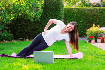 Beautiful young woman doing sport in garden outdoors following guide of online tutorial or trainer on laptop. Healthy lifestyle. Exercising at home. Doing fitness online