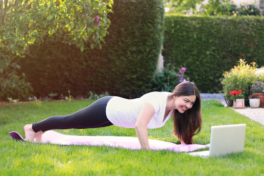 Beautiful young woman doing sport in her garden outdoors following guide of online tutorial or trainer on laptop. Healthy lifestyle. Exercising at home. Doing fitness online
