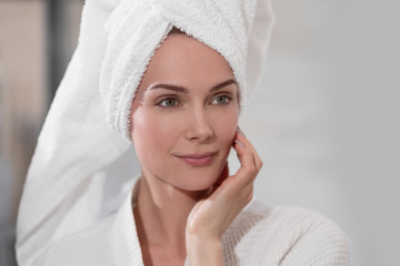 Attractive middle-aged blond woman with white towel on her head and in bathrobe standing in the bathroom by the mirror. She touches skin and smiles.