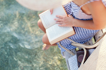 Woman is reading paper book on wooden pier at sea beach. Girl is enjoying summer vacation and...