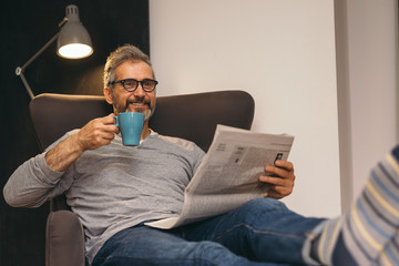 middle aged man relaxing in sofa reading newspaper and drinking tea at home