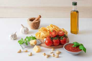 Table with ingredients to make tomato pesto. Tomatoes, garlic, fresh oregano and basil herbs, bottle of olive oil, few cashew nuts, parmesan cheese, cheese grater, wooden mortar and homemade pasta.