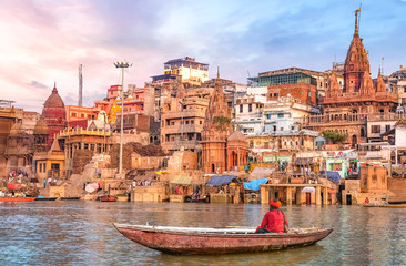 Ancient Varanasi city architecture at sunset with view of sadhu baba enjoying a boat ride on river Ganges. - obrazy, fototapety, plakaty