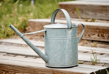 Old metal watering can standing on the wooden stairs of a terrace in the garden with grass and plants in the background - obrazy, fototapety, plakaty