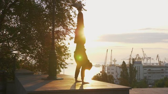 Silhouette of young man doing parkour handstand with sea port background during surise, slow motion