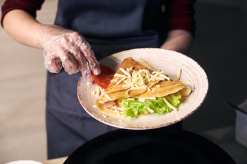Cropped view of chef decorate in white plate sandwich crepe with tomato slice, green salad and grated cheese. Food serving process. Appetizing hot stuffed dish with fish and vegetables. Close up view.