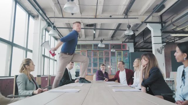 Manager start to dance on the table, sings into a megaphone, accelerates and slide on lap. Colleagues blow up flappers with confetti. Employees celebrate success. Corporate party business team