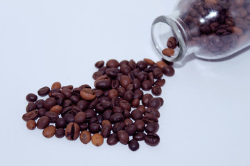Coffee beans are scattered in a heart shape from a transparent glass bottle on a white background