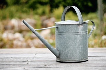 Old metal watering can standing on  wooden floor of a terrace in front of a beautiful springtime garden © franconiaphoto