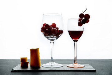 A few pieces of cheese and cherry tomatoes next to a glass of red wine filled with grapes on a white background