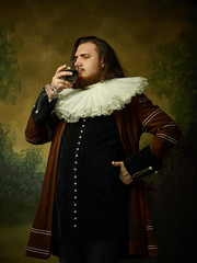 Young man as a medieval knight on dark studio background. Portrait in low key of male model in retro costume. Drinking red wine. Human emotions, comparison of eras and facial expressions concept.