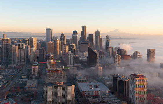 Seattle skyline with golden sunset light partially immersed in the fog in a cold autumn day with blue sky and the Mount Rainier visible on the background.