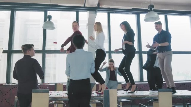 Employees celebrate the end of the project, have fun dancing on the table. Confetti fly around. Managers enjoy success and victory. Corporate party business team. Coworking