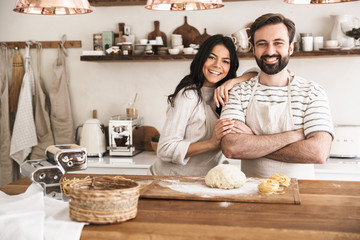 Portrait of joyful couple making homemade pasta of dough while cooking together in kitchen at home