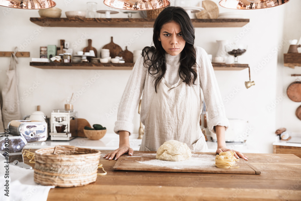 Wall mural portrait of irritated european woman making homemade pasta of dough in kitchen at home