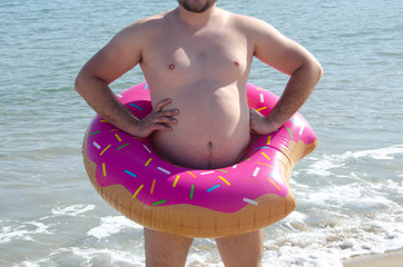 Fat man with obesity wearing inflatable donut ring on his belly on beach. Chubby man
