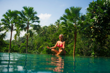 attractive and happy middle aged Asian Indonesian woman in bikini relaxed at infinity pool of exotic resort in tropical jungle sitting in lotus yoga position meditating