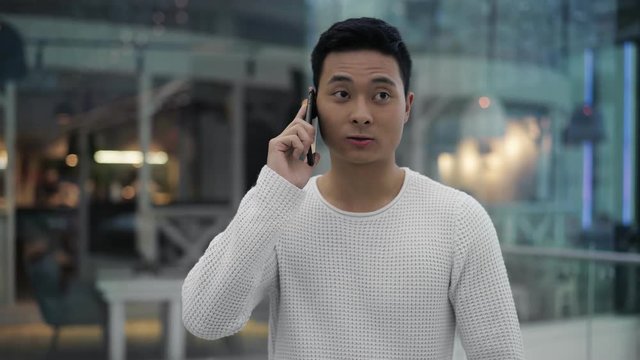 Middle shot of busy black hair asian male wearing white sweatshirt talking on phone while walking on background of glass windows in big mall center.