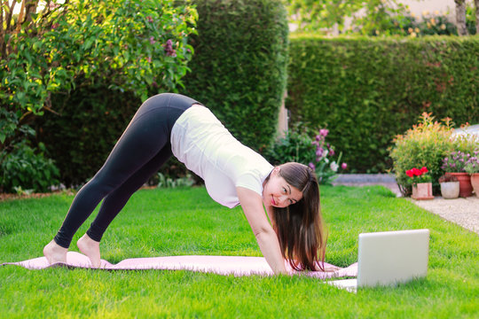 Beautiful young woman practicing yoga in garden outdoors following guide of online tutorial or trainer on laptop. Healthy lifestyle. Exercising at home. stretching muscles