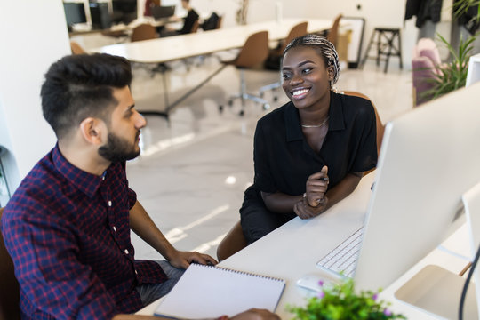 Successful Teamwork: Indian Business Man And African Woman Sitting At Desk Talking About Reports And Finance.