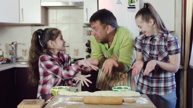 Funny father of the family, in a green kitchen apron jokes and has fun with his daughters while he teaches them to roll out the dough. Older girl with down syndrome.