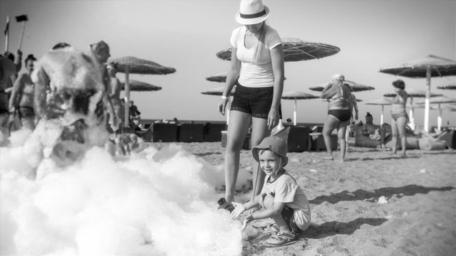Black and white photo of young woman with her toddler child son having fun and dancing on the soap foam beach disco party. Family relaxing on summer holiday vacation.