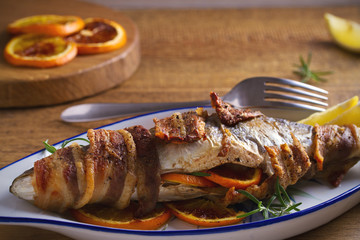 Grilled bacon-wrapped fish with oranges and rosemary, served with lemons - Image