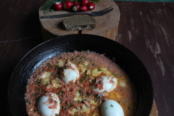 Black pan with stew eggs in tomato sauce on wooden background in rustic style
