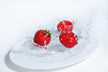 Fresh strawberries fall on a white plate. Splashes of water around the berries. White wooden table.
