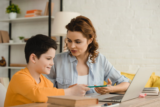 smiling mother helping adorable son doing homework while sitting at desk with notebook