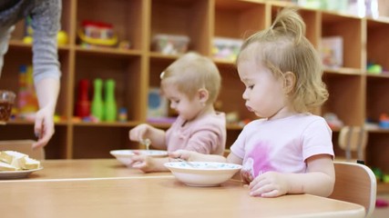 little kids are sitting at the table, they eat something from the plate by themselves using spoon, after that someone brings them drinks in mugs and puts them on the table, in the background is a