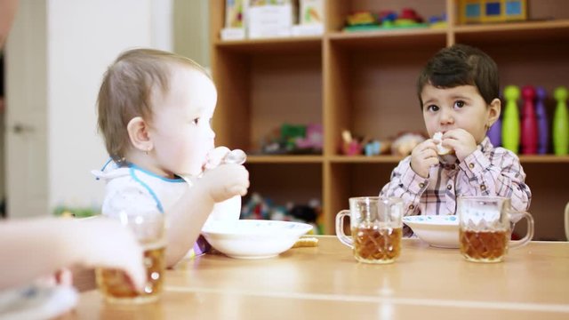 small children sit at the table, each one has a plate and a mug on the table , a little boy with dark hair eating bread and looking at the camera, on the background is cupboard with toys