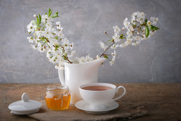 A cup of tea, a white kettle with cherry blossoms, a jar of honey on the table. Morning breakfast.