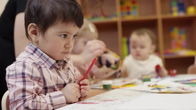 children sit at the table, the child in the close view holding a red marker, then puts it aside and reaches for something else, the children in the background paint with the teacher