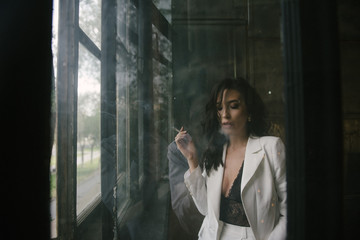 Young sexy brunette woman in suit smoking a cigarette by the window indoors