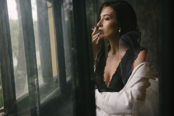 Fototapeta na wymiar Young sexy brunette woman in suit smoking a cigarette by the window indoors