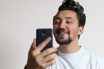 Portrait of a young man with braces smiling and looking at a smartphone. A happy young man admiring his braces on a white background