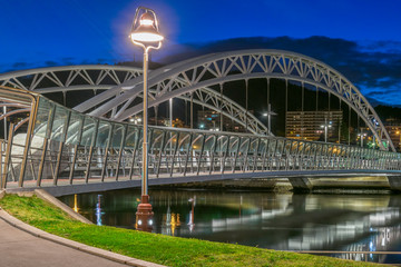 puente metálico blanco iluminado de noche con reflejo de las luces en el agua del rio con una farola en la vista