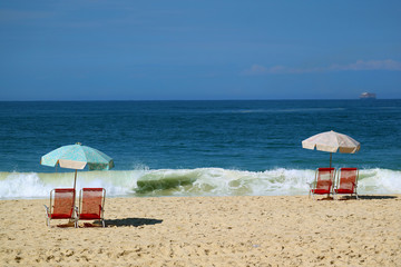 Two set of red beach chairs and a blue beach parasol on the sandy beach facing the crashing waves and vivid blue ocean