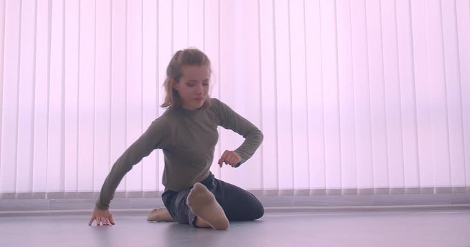 Tender caucasian professional ballerina warming up making leg-split in light studio on louvers background.