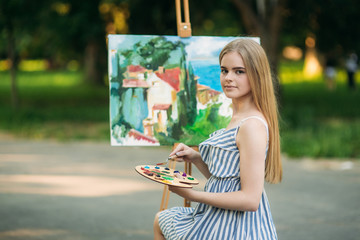 Beautiful blond hair girl sitting on stool and draws a picture in the park using a palette with...