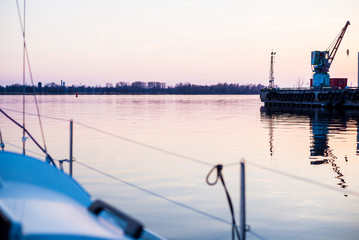 Close-up of the white yacht's bow. Sailing at sunset. Cargo ships and cranes in the background....