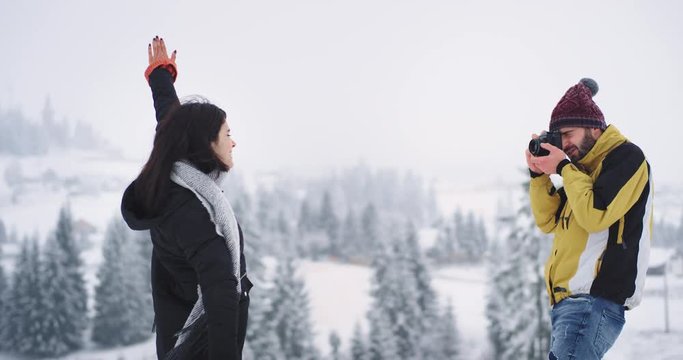 Positive young man and lady in amazing beautiful place on the top of snowy field , background beautiful snowy forest , man make pictures of the woman she funny posing in front of the photo camera
