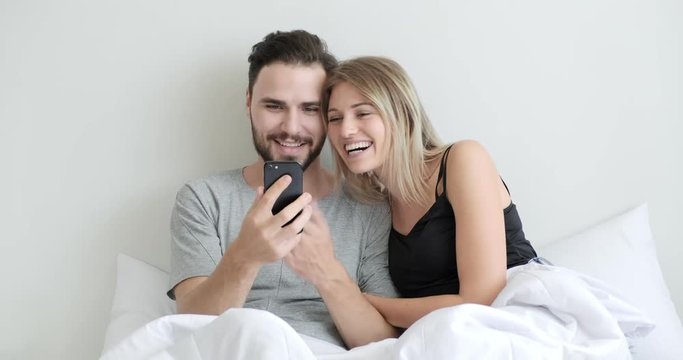 Couple using mobile phone to live stream, video call to friends in bed, surprise. Caucasian man and woman.